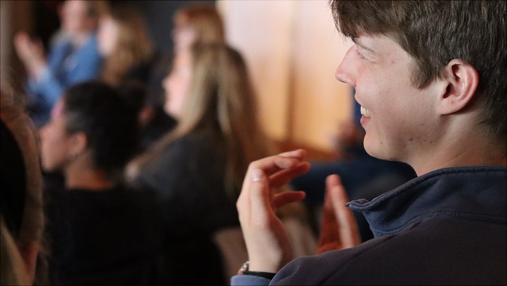 20240531-veranstaltung-im-jupiter-(c)-uhh,möller-(63)-733x414 Das Bild zeigt einen lächelnden jungen Mann, der in einer Gruppe von Menschen applaudiert.